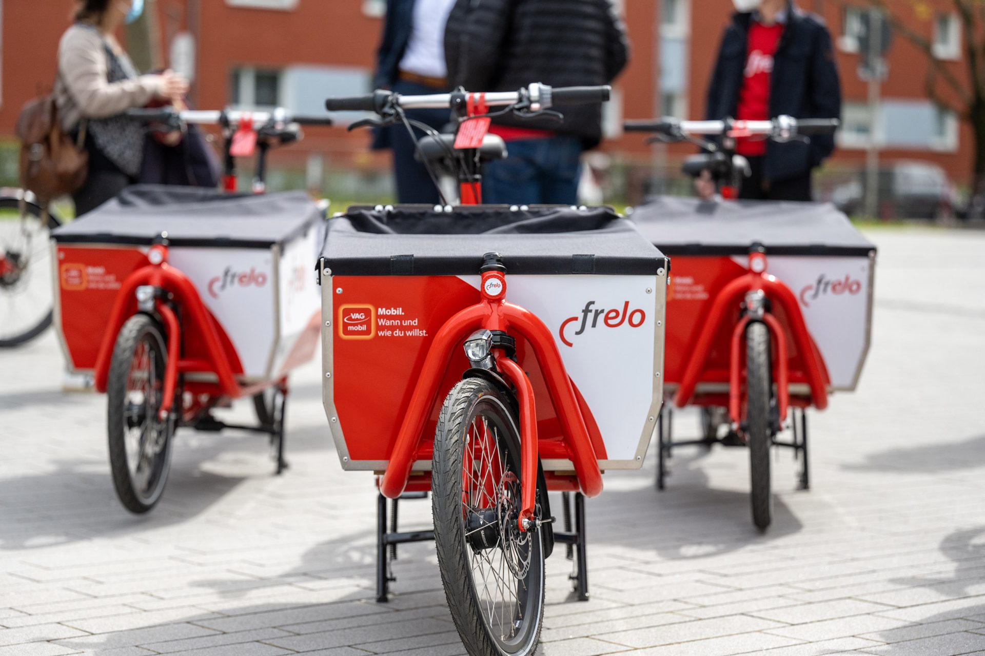 Drei rote frelo-Lastenräder. Im Hintergrund eine Gruppe Personen.